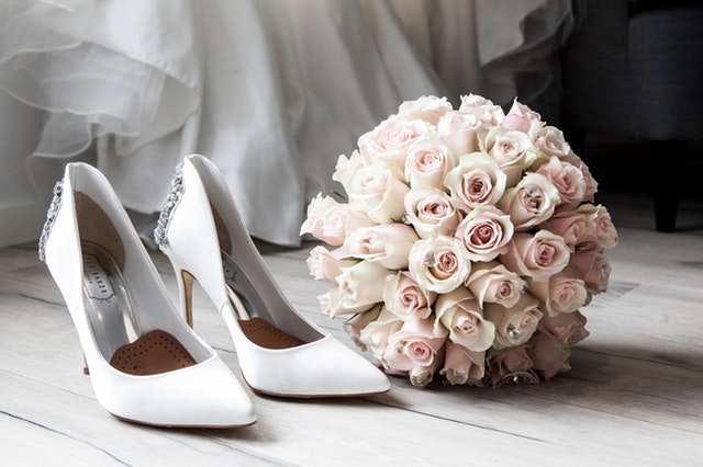 bílé lodičky, vedle svatební kytice z růží, za nimi kousek bílých šatů